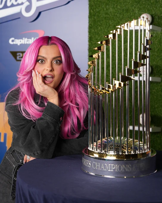 Bebe Rexha posing for a photo next to the World Series trophy. She has pink hair and is wearing a black jacket with black pants.