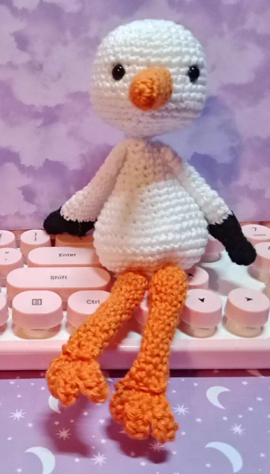 cigogne au crochet, exemple de présentation. le crochet est un art du fil qui permet  de réaliser ce genre de créations. La cigogne est assise sur un clavier d'ordinateur, le fond est un fond en papier cartoné avec des motifs de nuages. assise la cigogne mesure 12 cm environ. le fil utilisé est du coton