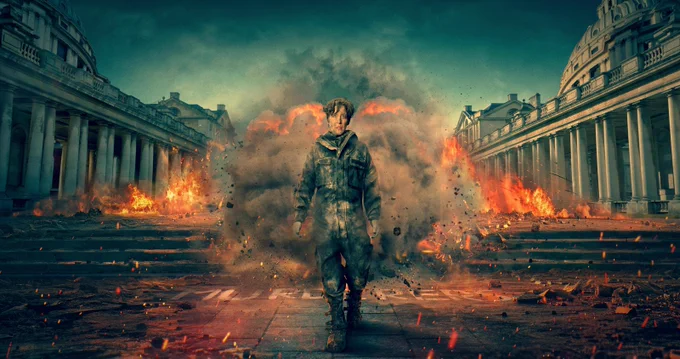 Vicky McClure in a dusty military-style uniform walks forward through a massive explosion between two rows of grand classical buildings with columns. Flames, smoke, and debris fill the air behind her, creating a dramatic, cinematic scene with intense lighting and destruction.