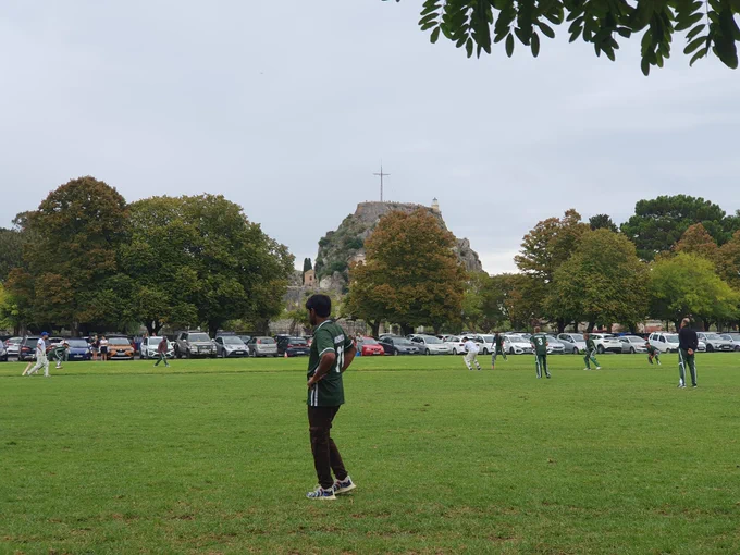 View from the boundary shows players in green and batters in white with old fortress in the background