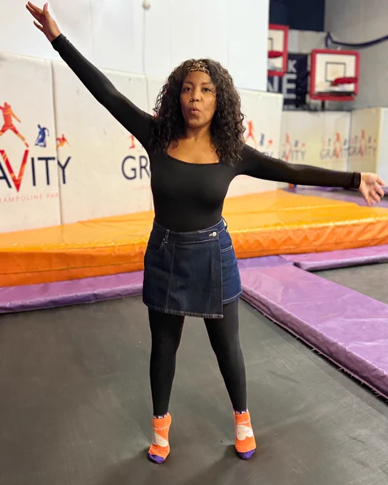 Person in a black top and denim skirt posing with one arm raised at a trampoline park named Gravity.