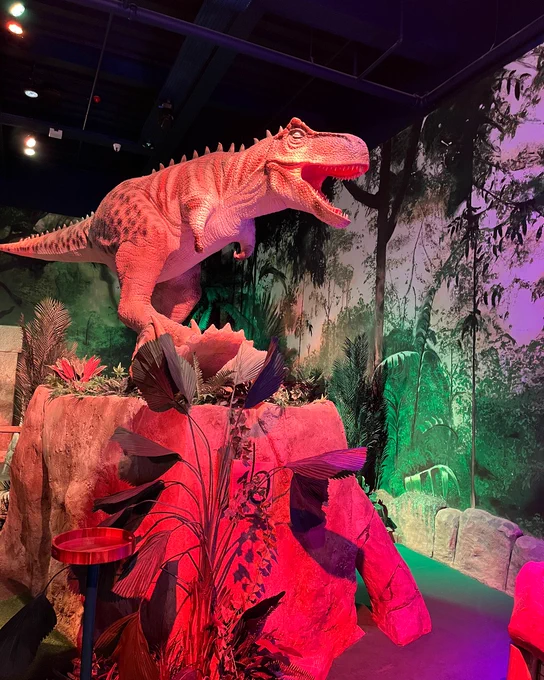 Display featuring a model of a Tyrannosaurus rex in a vibrant, jungle-themed exhibit with lush greenery and dramatic lighting.