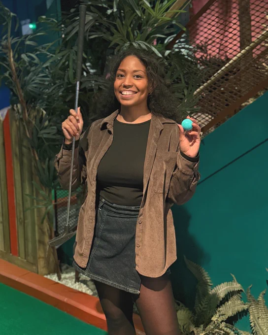 Person holding a golf putter and ball, smiling, standing on an indoor mini-golf course.