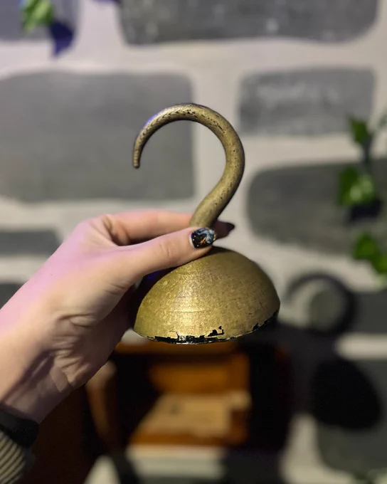 A hand holding a small, antique-looking gold bell with a curved top, in front of a blurred background featuring grey brick-like panels and green foliage.