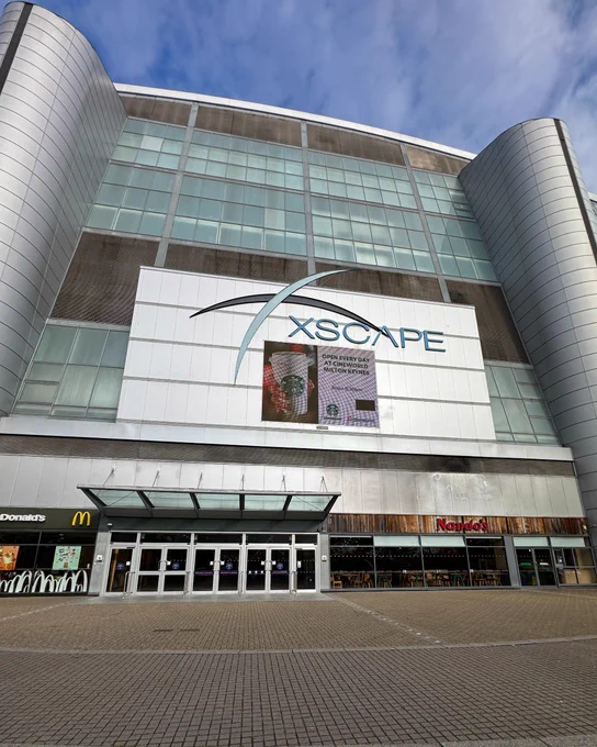 Exterior view of the Xscape entertainment complex, featuring a modern architectural design with prominent branding, alongside storefronts of McDonald's and Nando's at street level.