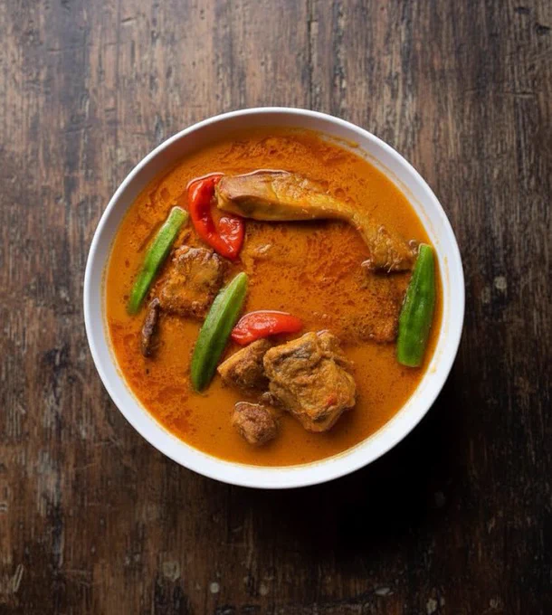 A high-angle close-up shot shows a white round bowl filled with a rich reddish-orange curry on a dark, distressed wooden surface. The curry looks thick and savory, with an oily sheen that catches the light.

Inside the bowl, a large piece of fish rests near the top center, partly submerged in the sauce. Around it are several dark chunks of cooked meat, likely mutton, covered by the curry. Three whole green okra pieces sit diagonally across the bowl, adding texture and contrast. Two bright red chili peppers bring a pop of color — one near the top center, another just below it.

The white bowl contrasts sharply with the dark wood background, whose rough grain and imperfections add a rustic feel. Focused lighting highlights the curry’s rich tones and texture, while the edges fade into shadow for a moody, dramatic look.