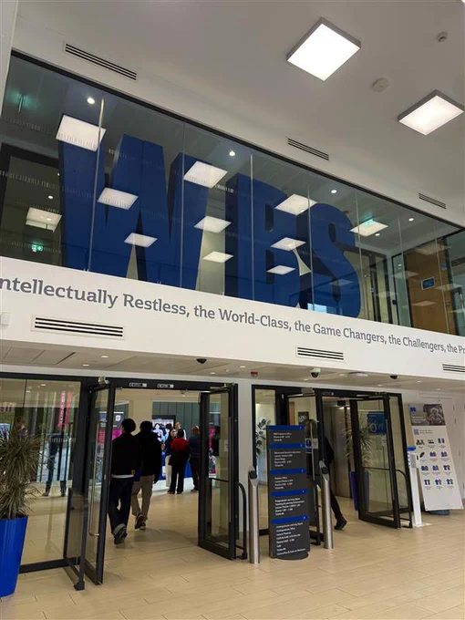 Entrance hall of Warwick Business School with large blue letters spelling 'WBS' above and people walking inside through glass doors.