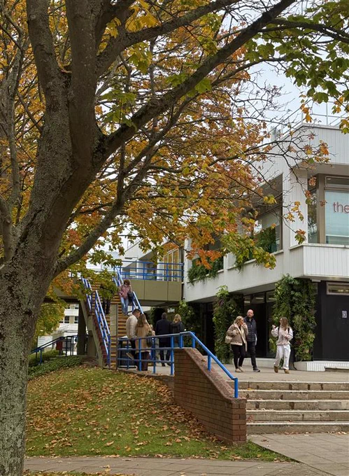 People walking and chatting as they walk past the University of Warwick library surrounded by autumn trees with orange and yellow leaves.