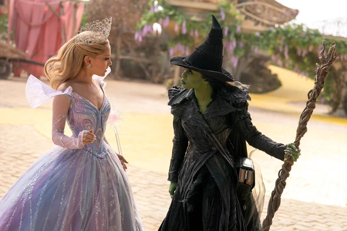 Ariana Grande as Glinda in a shimmering blue gown is confronted by Cynthia Erivo as Elphaba, holding a broomstick, in Munchkinland. The Yellow Brick Road can be seen behind them.