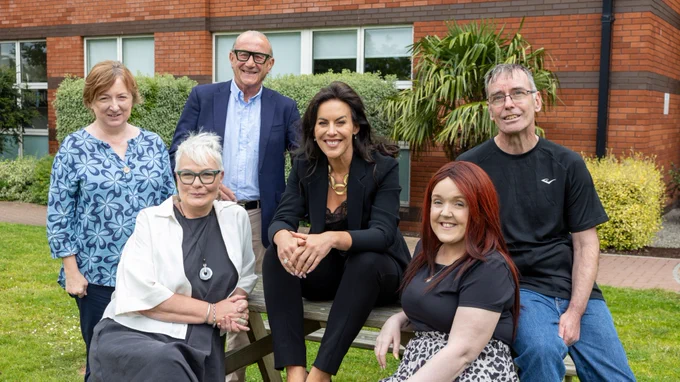 Group of six people posing outdoors on and around a bench, smiling, with a red brick building in the background. Photo: Paul Sherwood.