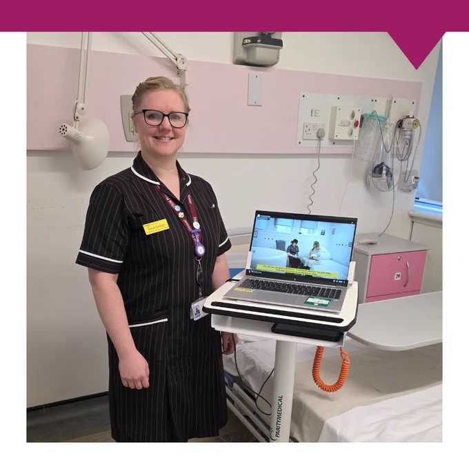 A healthcare professional in a black pinstripe uniform stands next to a mobile workstation in a hospital room. The workstation holds an open laptop displaying a paused video featuring two people seated in a clinical setting. The background includes a pink wall panel with medical equipment, sockets, and a mounted lamp. An orange coiled cable is attached to the workstation, and a hospital bed is partially visible on the right side of the image.