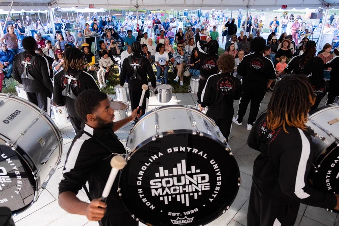 A group of young performers playing large drums under a tent, with focused expressions. They wear matching black jackets featuring the logo "Sound Machine" from North Carolina Central University. In the background, an audience sits and enjoys the performance, surrounded by a festive atmosphere.