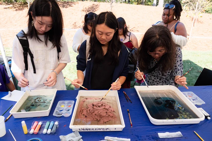 Three individuals are intently engaged in a creative activity at a table, working on water marbling with brushes and colored paints. They each have trays filled with vibrant paint mixtures in front of them. In the background, other participants can be seen observing or waiting to join the activity. The setting appears to be an outdoor event.