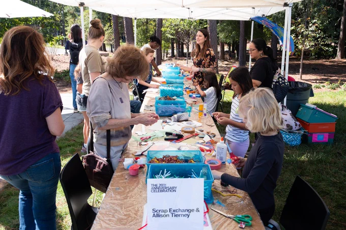 A group of people, including adults and children, are engaged in a creative craft activity at an outdoor table. Various materials are spread across the table, with participants working on individual projects. A sign reads "Scrap Exchange & Mission Team." Trees are visible in the background under a clear sky.