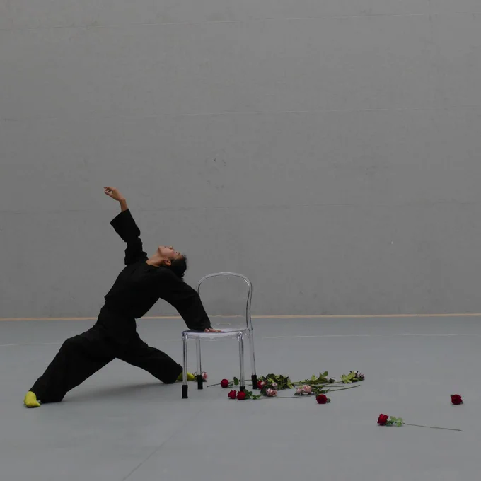 Dancer in black outfit stretching backwards with one hand on a transparent chair surrounded by scattered roses on a grey floor.