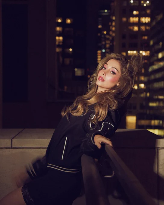 Woman on the top of a building at night wearing a varsity jacket, the background is an NYC cityscape, the buildings lit up just out of focus. The woman is posing and smiling.