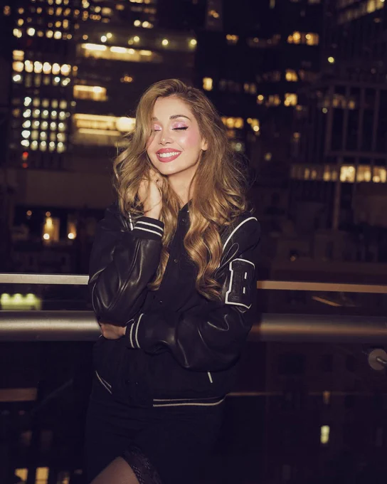 Woman on the top of a building at night wearing a varsity jacket, the background is an NYC cityscape, the buildings lit up just out of focus. The woman is posing and smiling.