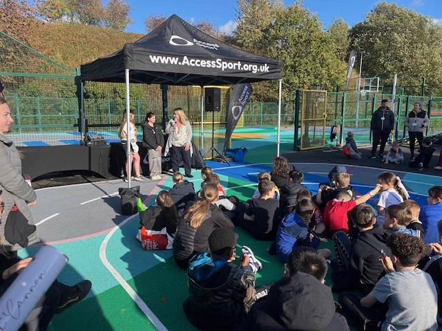 Photo of Eileen Buchan from Salford Youth Service and 2 young people doing a speech. They are stood under a gazebo with children sat listening to them.