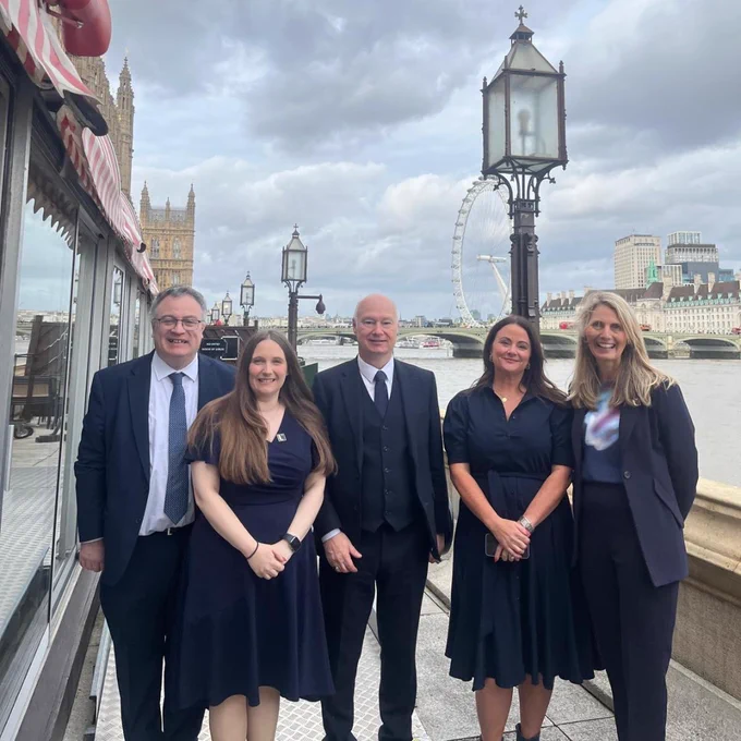 A group of five people—three women and two men—stand for a photo outdoors in London. The background features a large historic lamp post, the River Thames, the London Eye, and the Houses of Parliament.