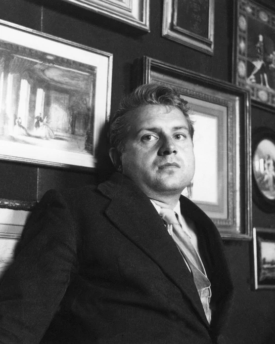 Image description: The artist Francis Bacon is lent against a wall. The wall is covered in framed paintings and drawings. He is wearing a dark jacket and a light shirt and tie.