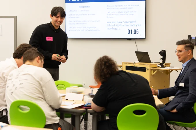 Four individuals participate in a workshop or class setting with a digital timer reading 1:05 displayed prominently on a screen in the background. One person stands with papers, presenting to the group while the others are seated, engaging with various documents at tables.