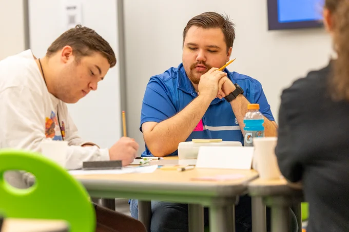 Three individuals engaged in a discussion at a table with notebooks and a drink visible. One person is in deep thought, holding a pencil to their chin.