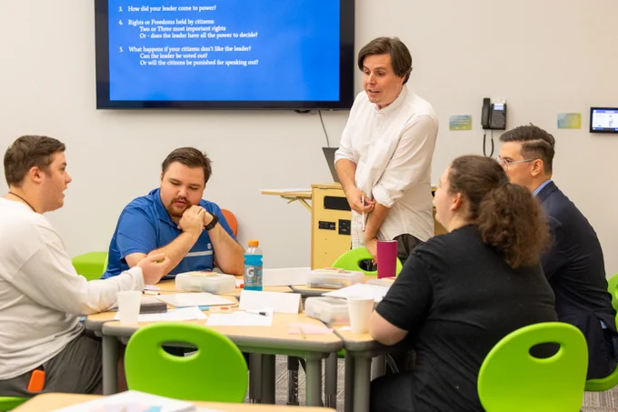 A group of six adults engaged in a discussion around a table in a brightly lit classroom. A whiteboard lists discussion points in the background.