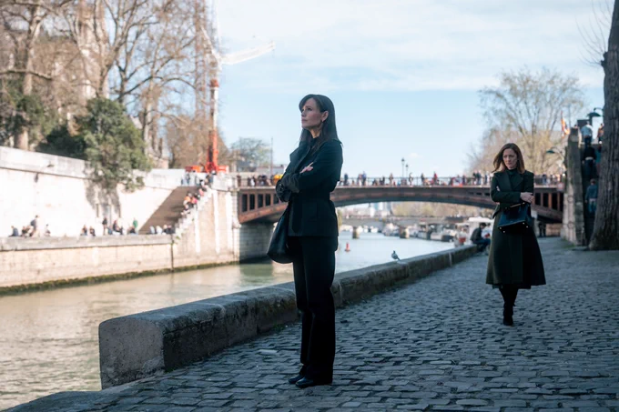 From The Last Thing He Told Me. Hannah Hall portrayed by Jennifer Garner stands on a rocky path by a body of water. Judy Greer’s character is walking towards her.