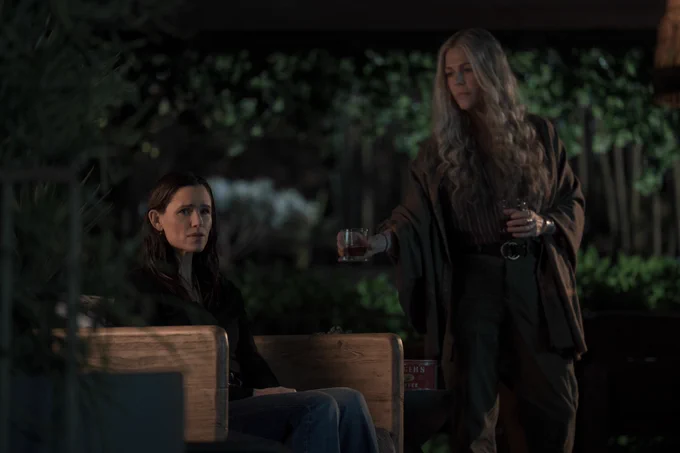 From The Last Thing He Told Me. Rita Wilson’s character is standing and holding two glasses leaning to give one to Hannah Hall portrayed by Jennifer Garner who is sitting on a chair looking distressed.