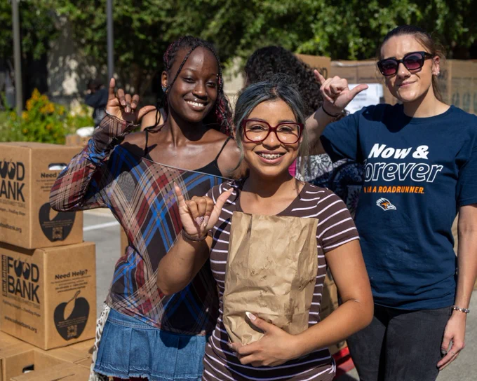 Three volunteers smiling at a Roadrunner Distribution event, standing in front of boxes labeled 'Food Bank.' The person in the middle holds a brown paper bag. The three people are holding up a "Birds Up" hand gestor. 