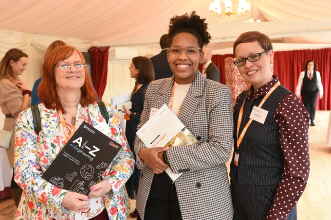 Three professionals at the Engineers 2030 parliamentary event on National Engineering Day  holding a book titled "AI-Z of Engineering," smiling for the camera in a tented venue.