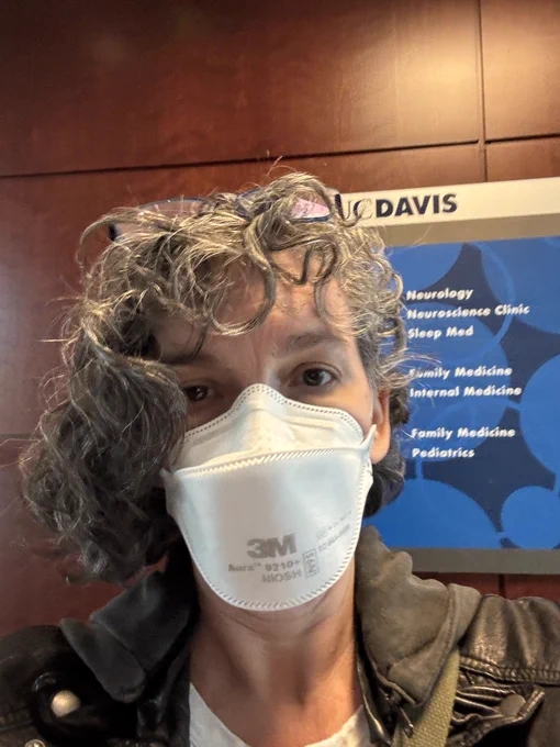 A middle aged white woman is wearing a 3m aura mask in front of a UC Davis medical sign.