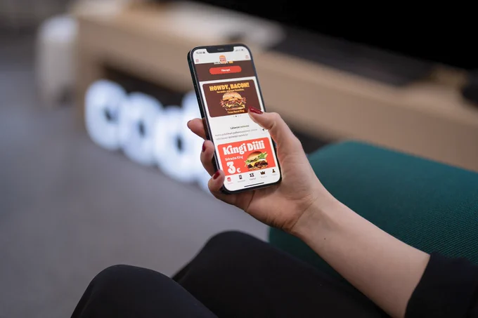 A hand holding a phone where Burger King Application is open. In the app there's a menu selection and promoted burger deals. In the background, there's a tv stand, tv and Codemate logo blurred.