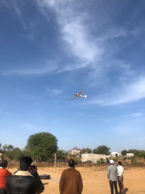 Group of men watching and photographing a small drone flying in a clear blue sky over a rural area with trees and buildings.