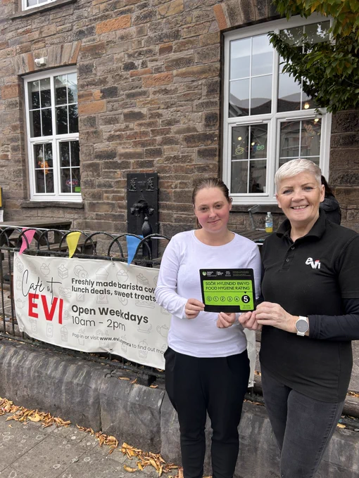 Two women smile at the camera holding a green '5' hygiene rating certificate, next to a Cafe at EVI banner on the railings outside the building. 