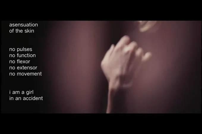 A still from a film. A hand is the foreground. Words are to the left of the screen: asensuation of the skin, no pulses, no function, no flexor, no extensor, no movement. I am a girl, in an accident. 