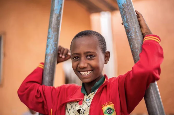 A joyful child in a red Brazil jacket smiles while holding onto a metal ladder outdoors.