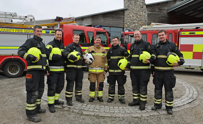 Six firefighters, pose smiling with their Assistant Chief Fire Officer to the centre, in full fire kit, helmets under arm, in front of two fire engines.