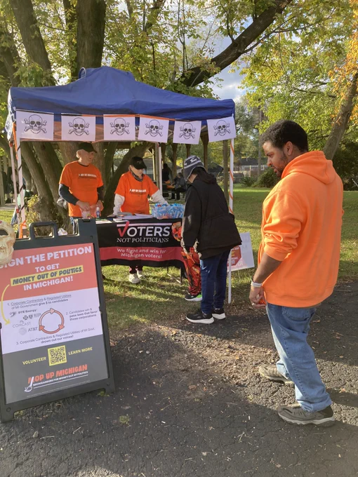 Voters Not Politicians signature collection table halloween decorated with skulls, someone reading a sign - "Sign The Petition to get Money Out Of Politics in Michigan!" 