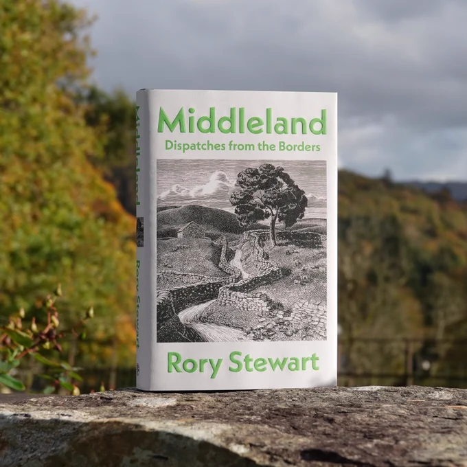 Rory Stewart, Middleland: Dispatches from the Borders (Jonathan Cape) standing on a stone wall in Grasmere with autumn trees, fell and grey sky beyond