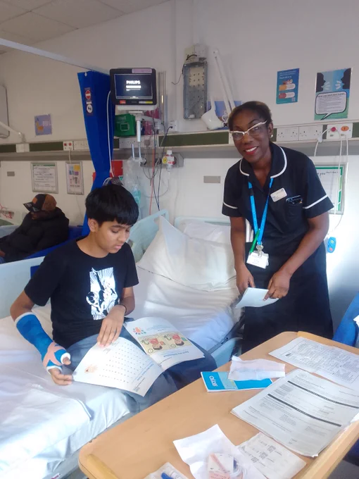 A young patient with a blue cast on his arm reads an activity booklet while sitting on a hospital bed, attended by a nurse holding papers.