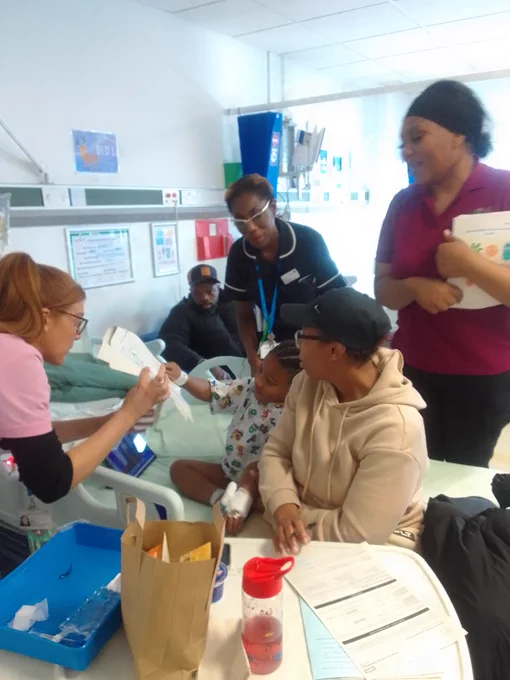 A child in a hospital bed receives an activity booklet from a healthcare worker while family members watch nearby in a bright hospital room.