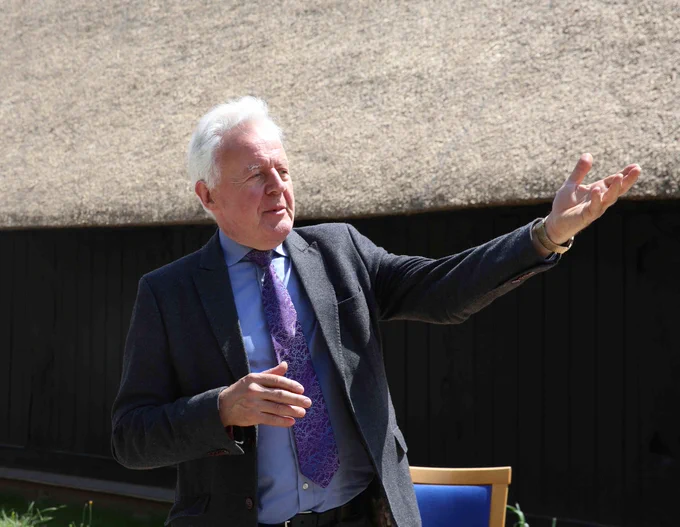 A photograph of a man standing outdoors, gesturing with his right hand in front of a thatched roof and a wooden structure. 
