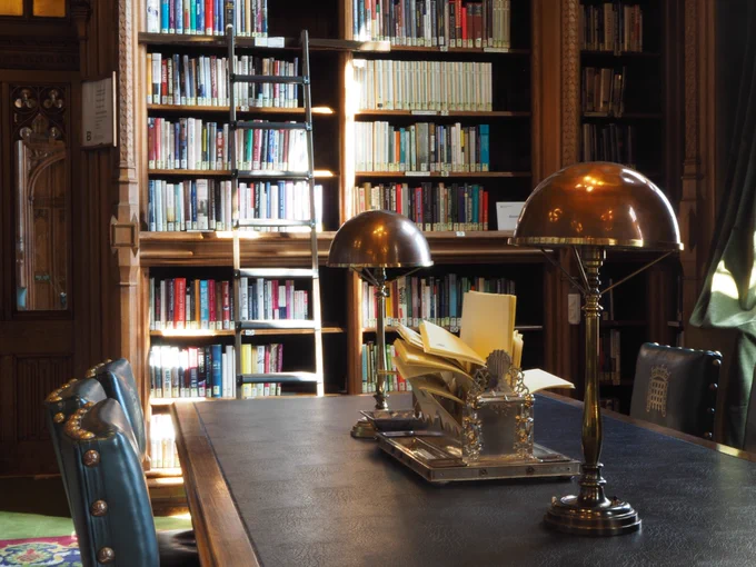 A desk in the Commons Library surrounded by green leather chairs. On top of the desk are two brass lamps  and an ornate silver stationary organiser filled with paper. In the background are bookshelves with a ladder resting against them.