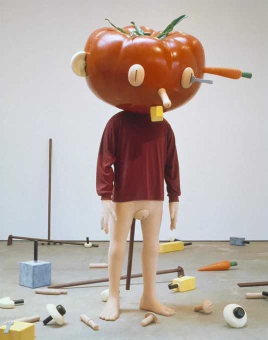 A beige doll wearing a burgundy sweater and a plastic tomato head is standing in a white and grey room scattered with doll parts and tools.