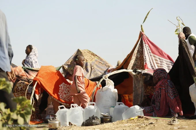 Children and families sit outside tents with water cans in a camp for the displaced