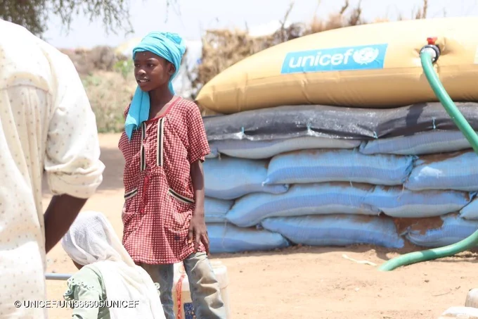 A girl stands next to other adults in a camp for displaced, with UNICEF supplies in the background