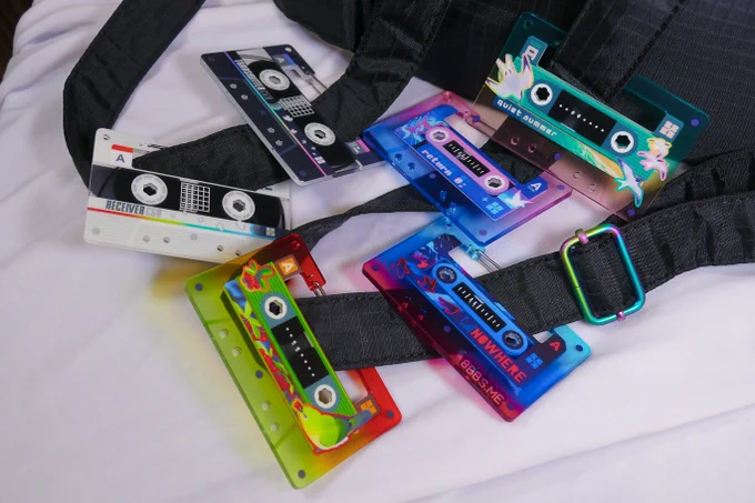 A collection of 6 acrylic carabiners hooked onto the straps of a small black backpack. They are colourful and feature bird-themed designs on cassette-tape shapes.