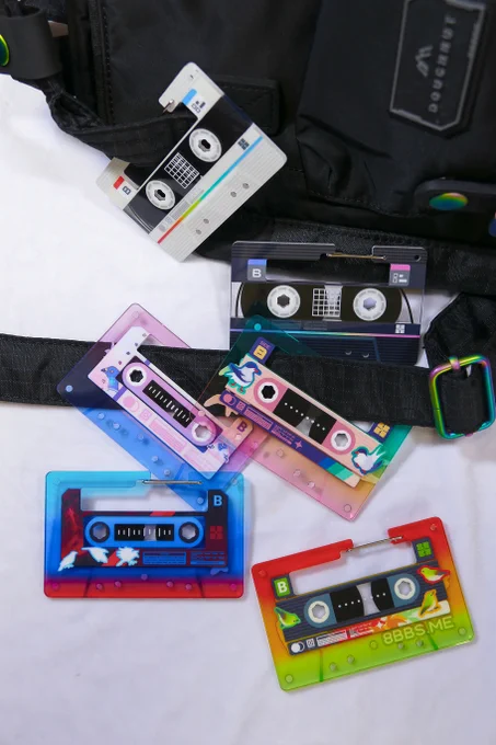 A collection of 6 acrylic carabiners hooked onto the straps of a small black backpack. They are colourful and feature bird-themed designs on cassette-tape shapes.
