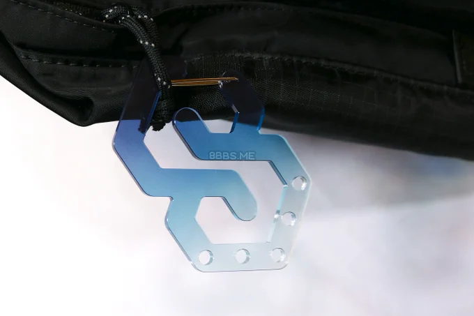 An acrylic carabiner in the shape of the cool S, coloured blue gradienting to clear at the bottom, where there are 5 holes for charm attachment. It is hooked onto the zipper pull of a black bag.